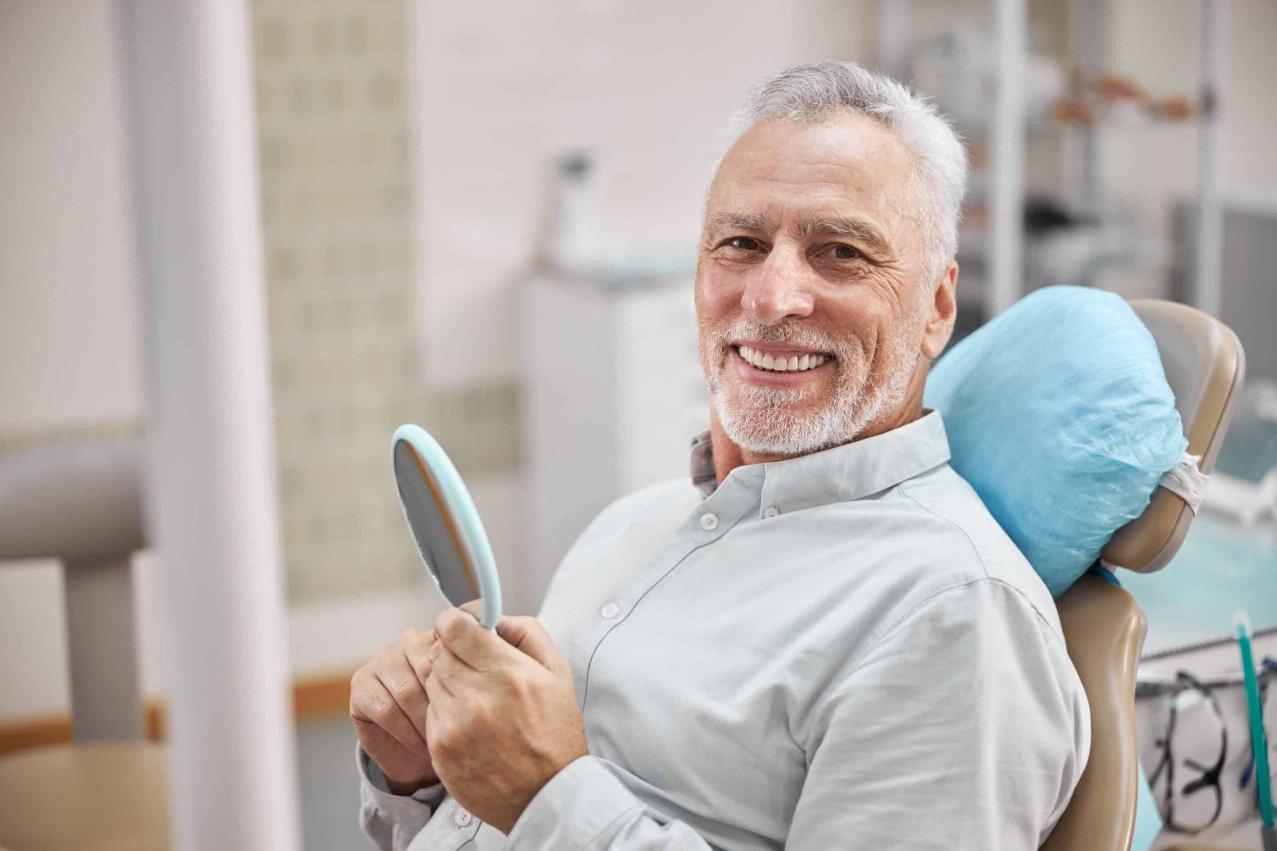 Cheerful patient sitting in a dental chair and holding a mirror while smiling at the camera.